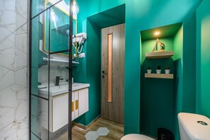 Un baño con una pared verde, una puerta de madera, un lavamanos blanco con detalles dorados y una estantería con plantas.
