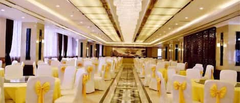 Salão de banquetes com fileiras de mesas e cadeiras, um corredor central e um candelabro.
