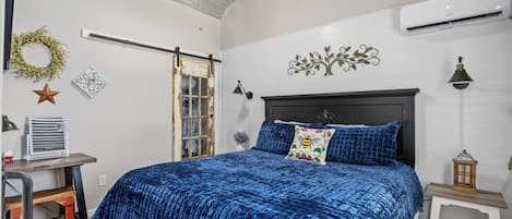 Un dormitorio con cama, escritorio, silla, una mesita, un calentador de pared, ventilador de techo, ventana y un adorno decorativo.