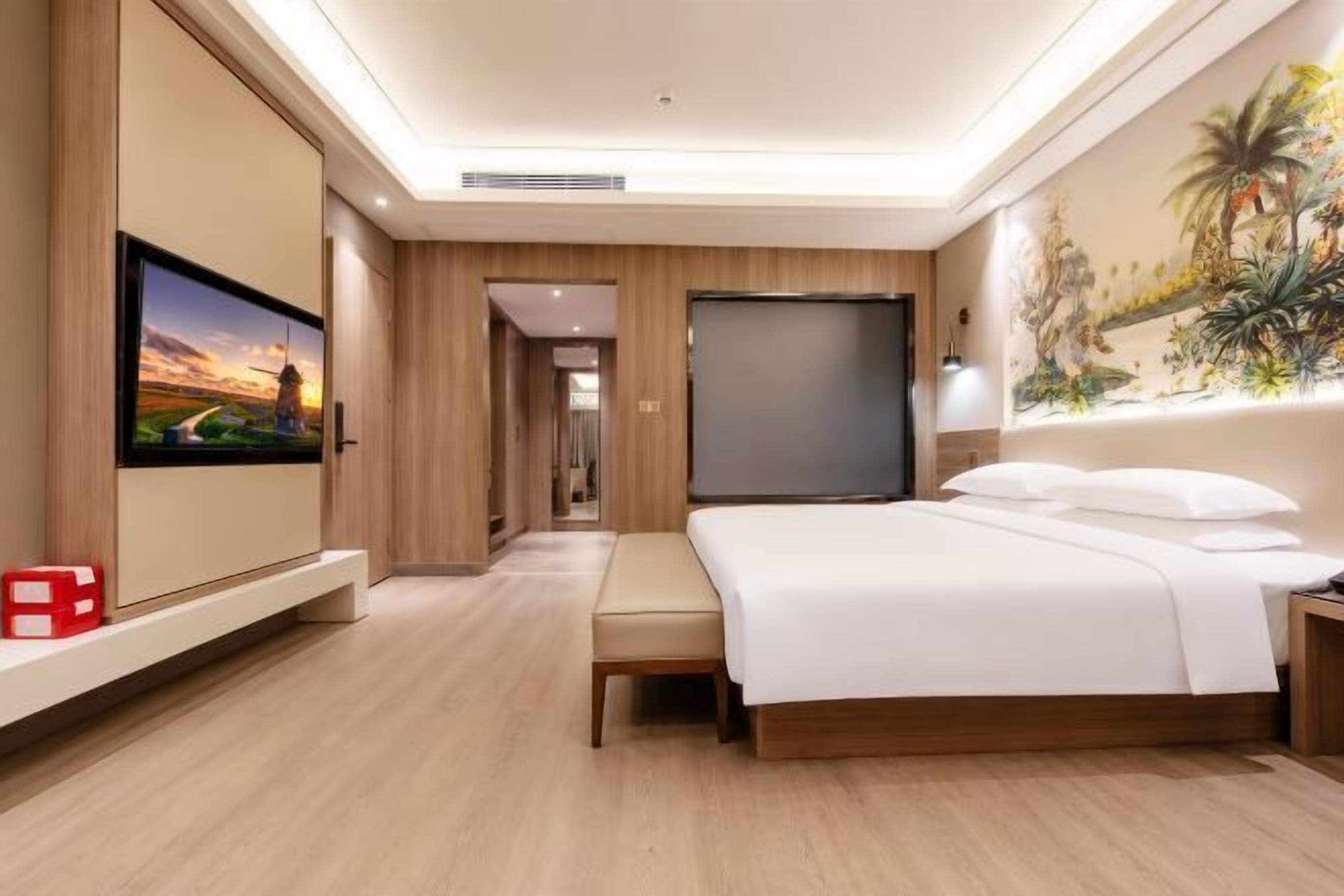 A hotel room with a bed, a TV, a painting, and a hallway leading to another room.