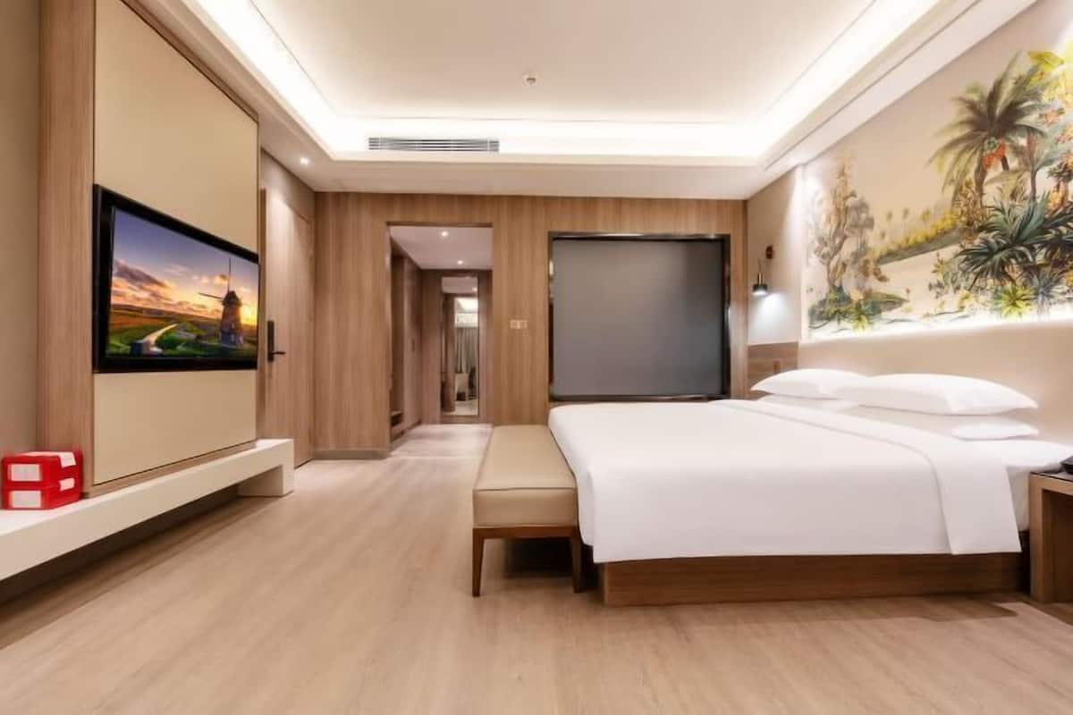 A hotel room with a bed, a TV, a painting, and a hallway leading to another room.