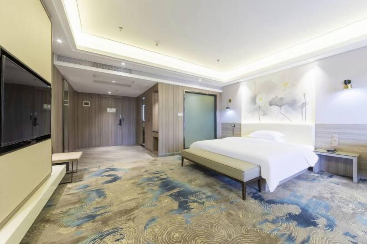 A modern hotel room with a large bed, a flat-screen TV, a bench, and a wall-mounted lamp.