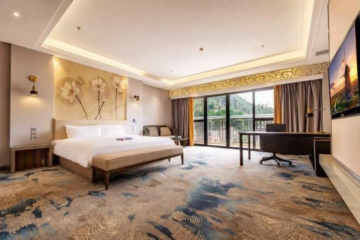 A hotel room with a large bed, a desk, a chair, a balcony with curtains, and a wall-mounted artwork.