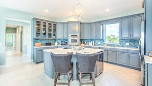 Une cuisine moderne avec des meubles gris, un îlot central doté de deux tabourets de bar, un lustre et une vue sur l’extérieur.