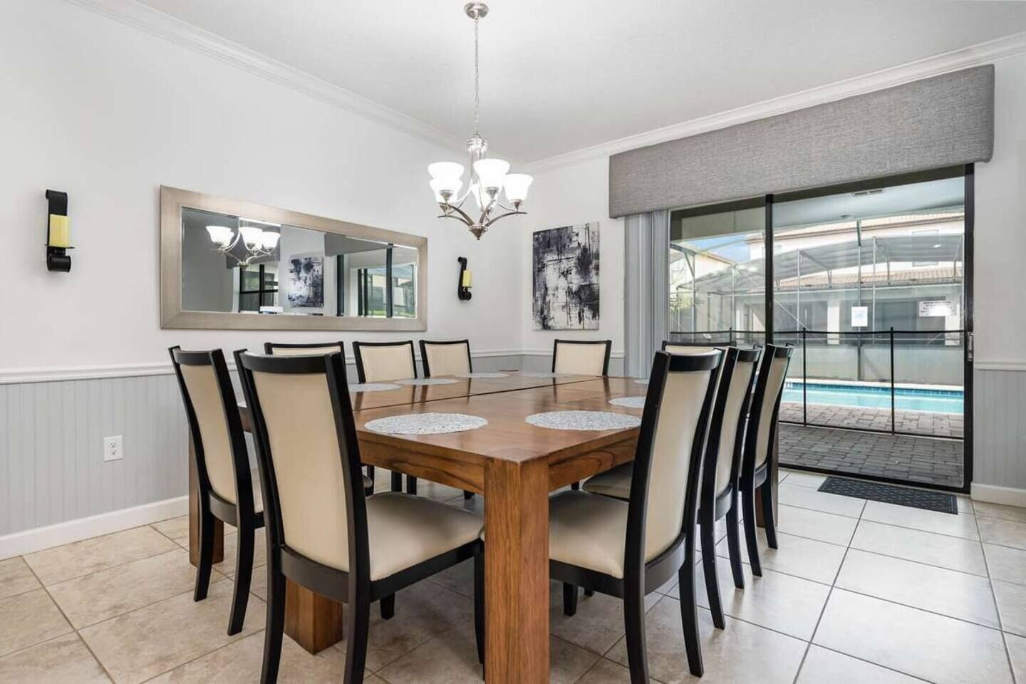 A dining room with a large wooden table, six chairs, a chandelier, a large mirror, and a view of a swimming pool through a sliding glass door.