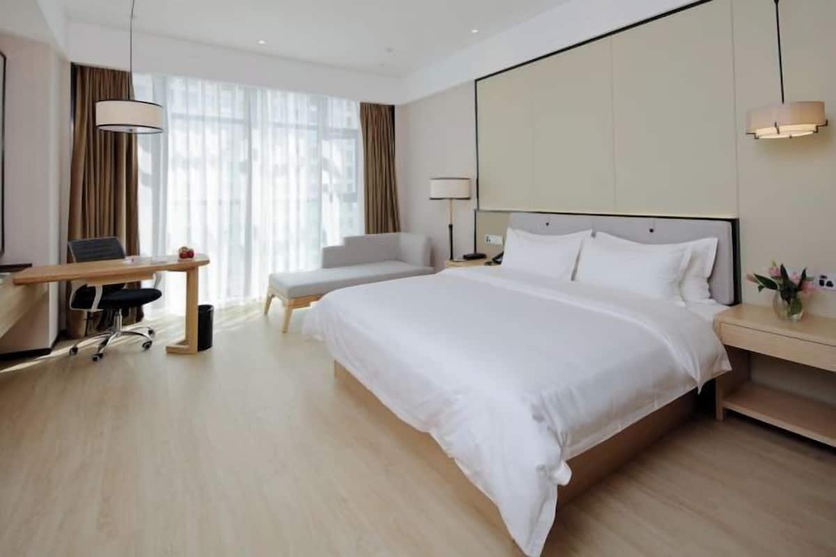 A modern hotel room with a large bed, a desk with a chair, a sofa, and a large window with curtains.