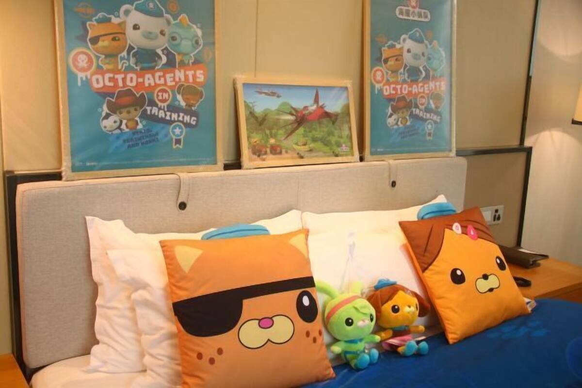 Een hotelkamer met een bed met meerdere kussens, waaronder een paar met tekenfilmfiguren. Achter het bed hangen posters aan de muur.