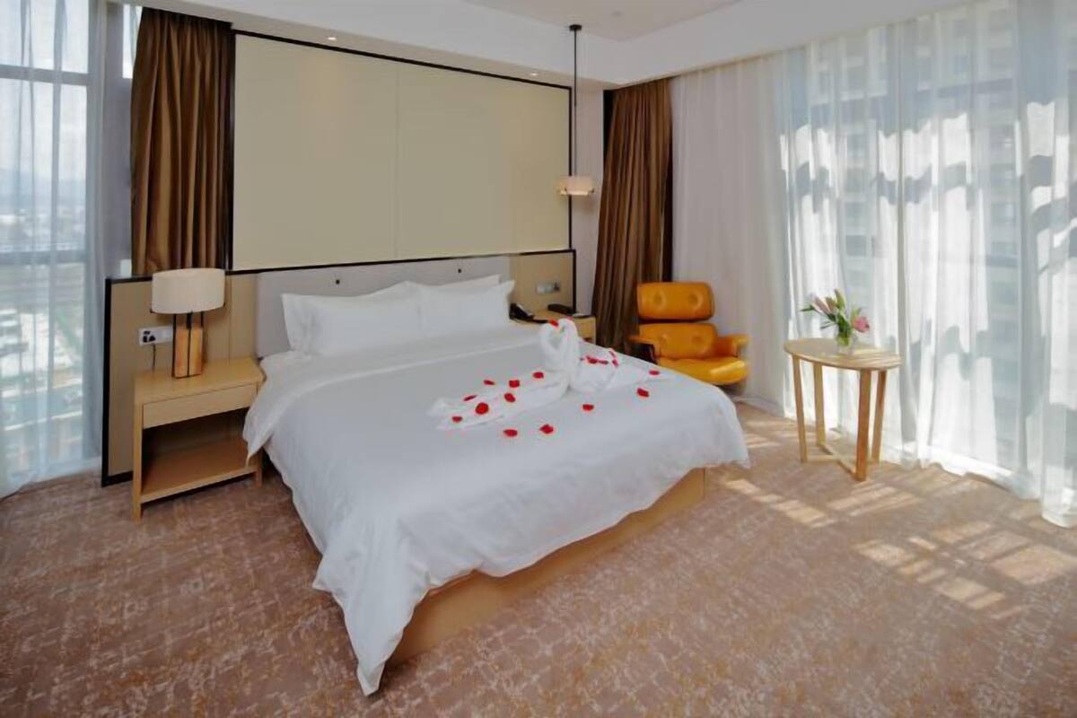Een hotelkamer met een groot bed, nachtkastje, lamp, stoel en een tafeltje met een vaas.