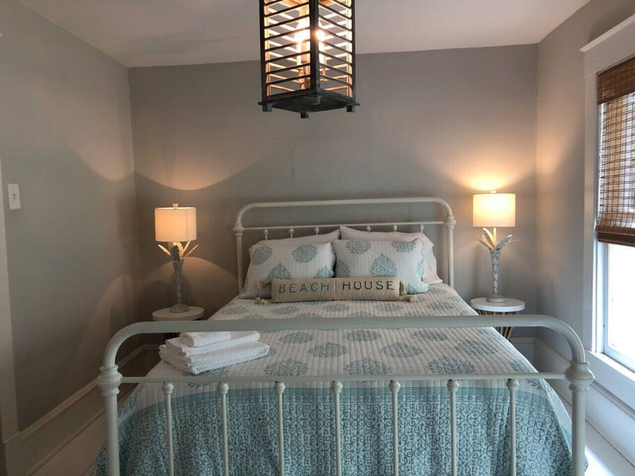 Cama arrumada com travesseiro "BEACH HOUSE", duas luminárias de cabeceira e um lustre em quarto com paredes cinza claro.
