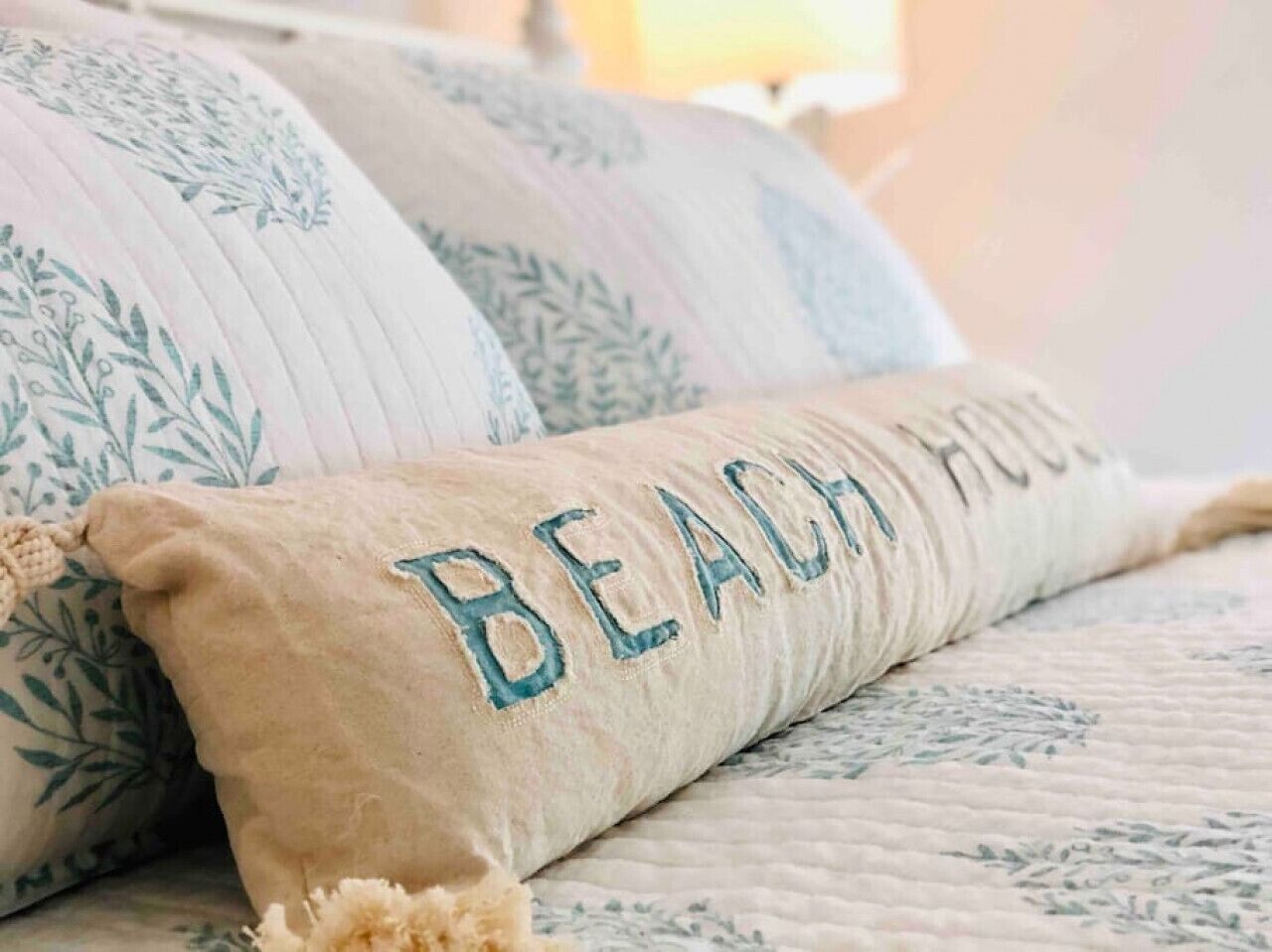 A bed with a beige pillow and a pillowcase with "BEACH HOUSE" embroidered on it.