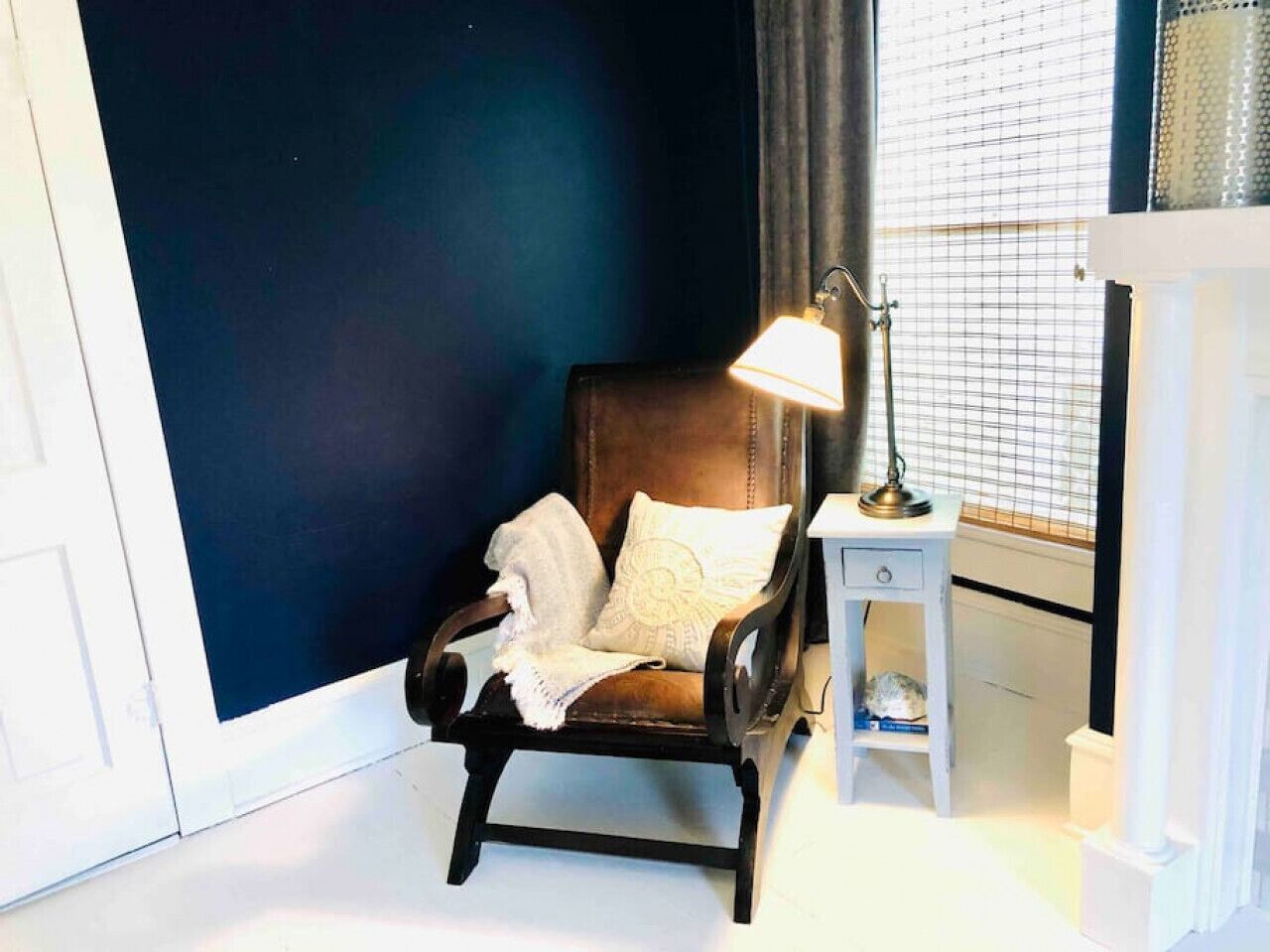 A room with a dark blue wall, a wooden armchair with a white throw blanket and pillow, a small white nightstand with a lamp, and a window with blinds.