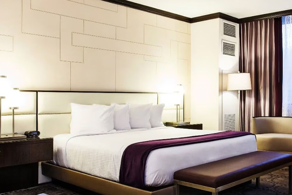 A hotel room with a bed, bedside tables, a bench, a chair, and a large window with curtains.