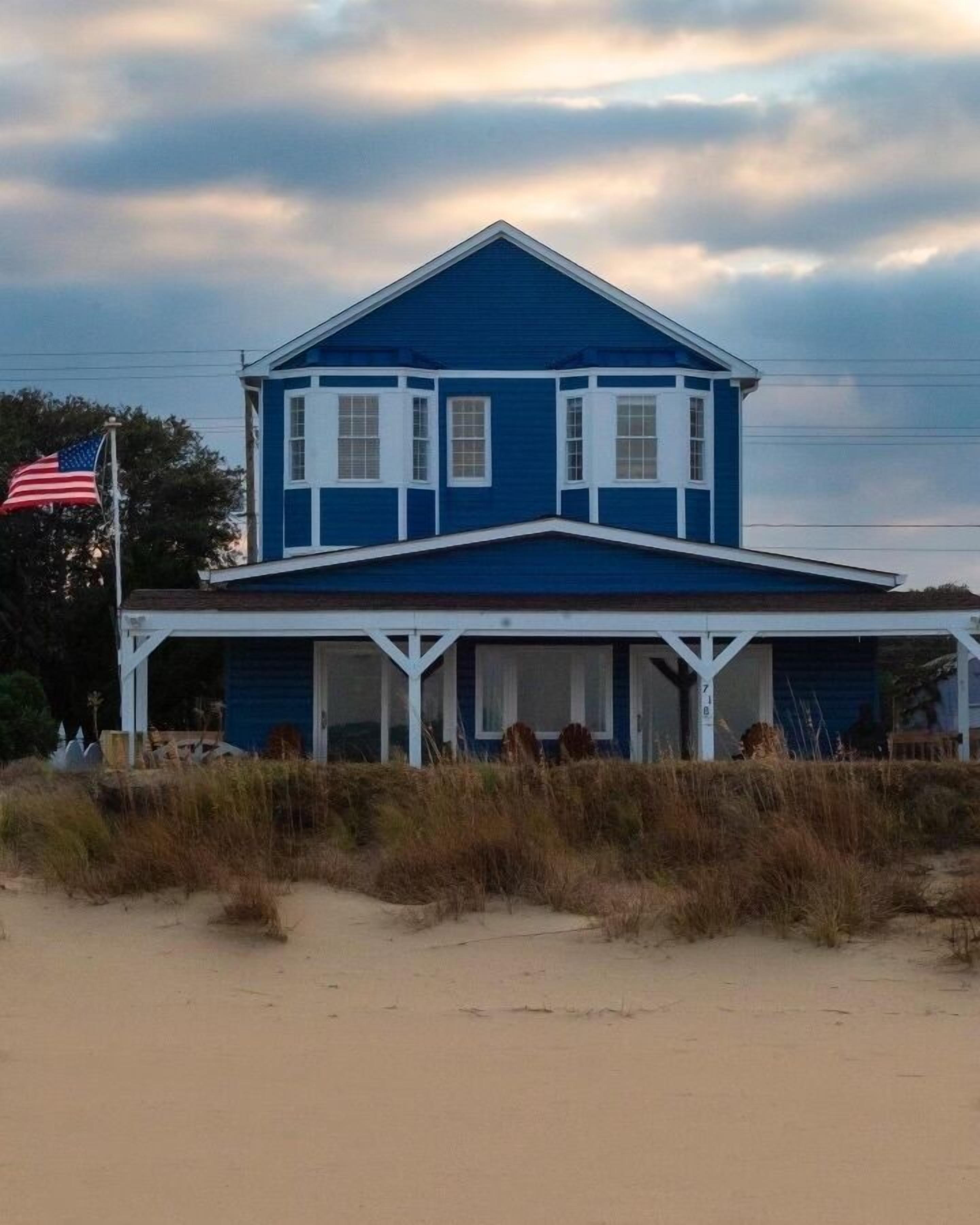 A two-story house with a blue exterior and white trim, featuring a covered porch and a flag on the left.