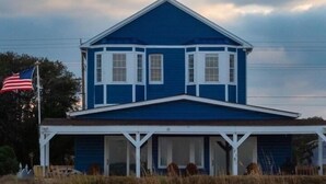 A two-story house with a blue exterior and white trim, featuring a covered porch and a flag on the left. - Romantic Beachfront Cottage 🐕 💙 (Norfolk)