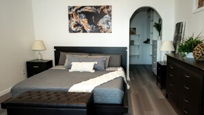 A modern bedroom with a large bed, a dark wooden dresser, and a nightstand with a lamp. - Romantic Beachfront Cottage 🐕 💙 (Norfolk)