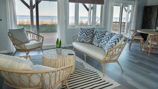 A spacious living area with wicker furniture, a round coffee table, and a view of the beach. - Romantic Beachfront Cottage 🐕 💙 (Norfolk)
