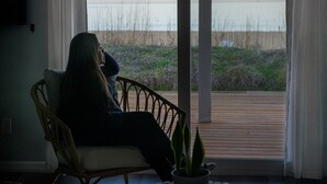 A person sitting on a wicker chair by a window overlooking a wooden deck and a beach. - Romantic Beachfront Cottage 🐕 💙 (Norfolk)