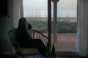 A person sitting on a wicker chair by a window overlooking a wooden deck and a beach.