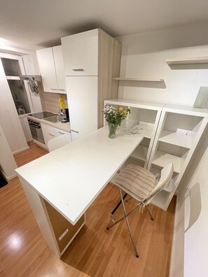 A compact kitchen with white cabinets, a built-in oven, and a small dining area with a foldable chair. - Schönes Appartment in Laufweite zum Zentrum & Mainufer (Würzburg)