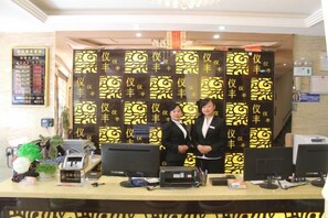 A reception desk with two individuals standing behind it, a computer, and a wall with repeated patterns. - Yi Feng Bussiness Hotel (Anning)