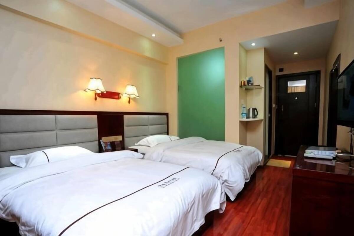 Habitación de hotel con dos camas, cabeceros, lámparas de noche, pared verde, una puerta y un televisor.