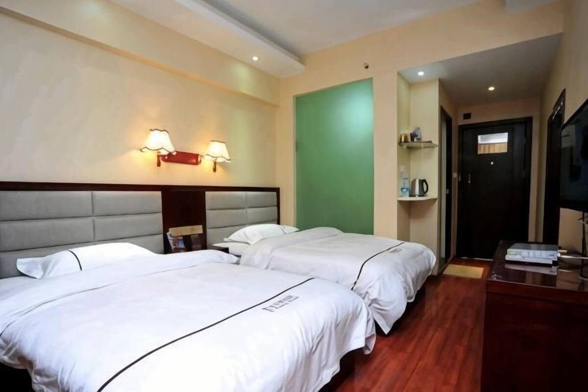 Habitación de hotel con dos camas, cabecera, lámparas fijadas a la pared, pared de acento verde, suelo de madera, escritorio con silla y televisión.