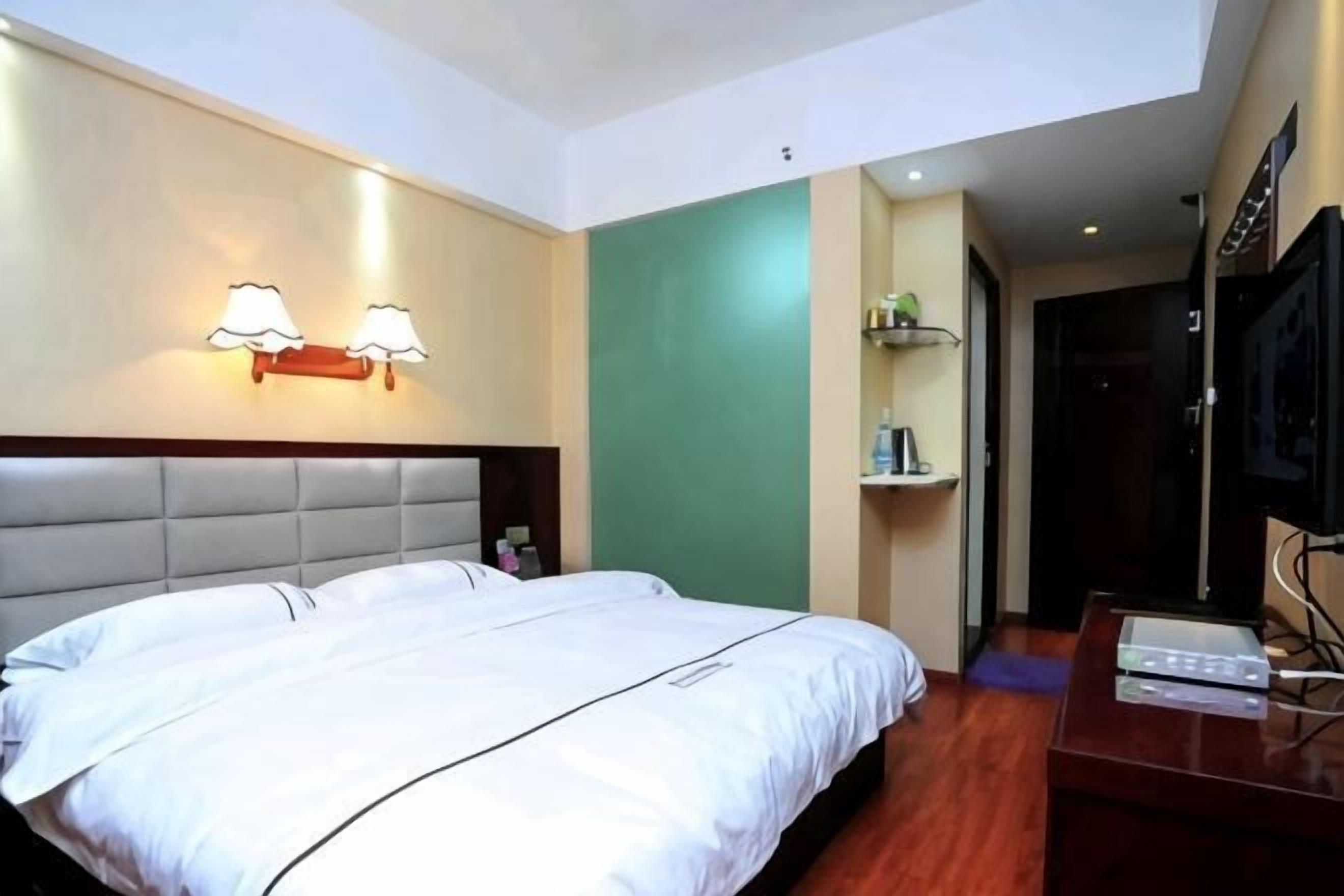 A hotel room with a large bed, a headboard, a wall-mounted light fixture, a television, and a wooden floor.