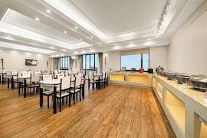 Un comedor amplio con múltiples mesas y sillas, una barra de buffet con recipientes de comida, y grandes ventanales que ofrecen vistas.