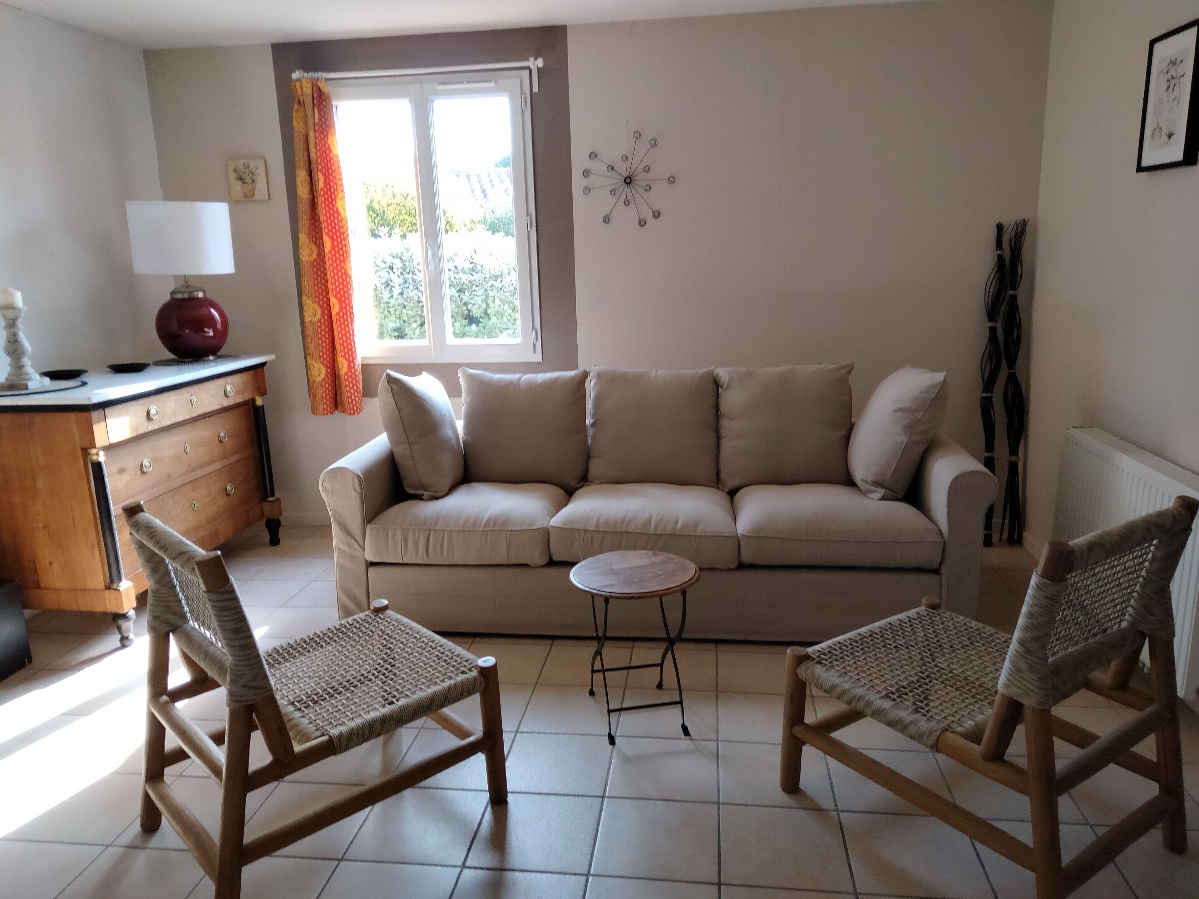 A living room with a sofa, two wicker chairs, a small round table, a lamp, a radiator, and a window with curtains.