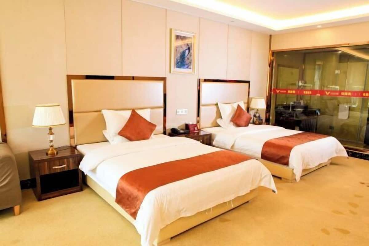 A hotel room with two beds, a nightstand with a lamp, a wall-mounted artwork, and a large window with a view.