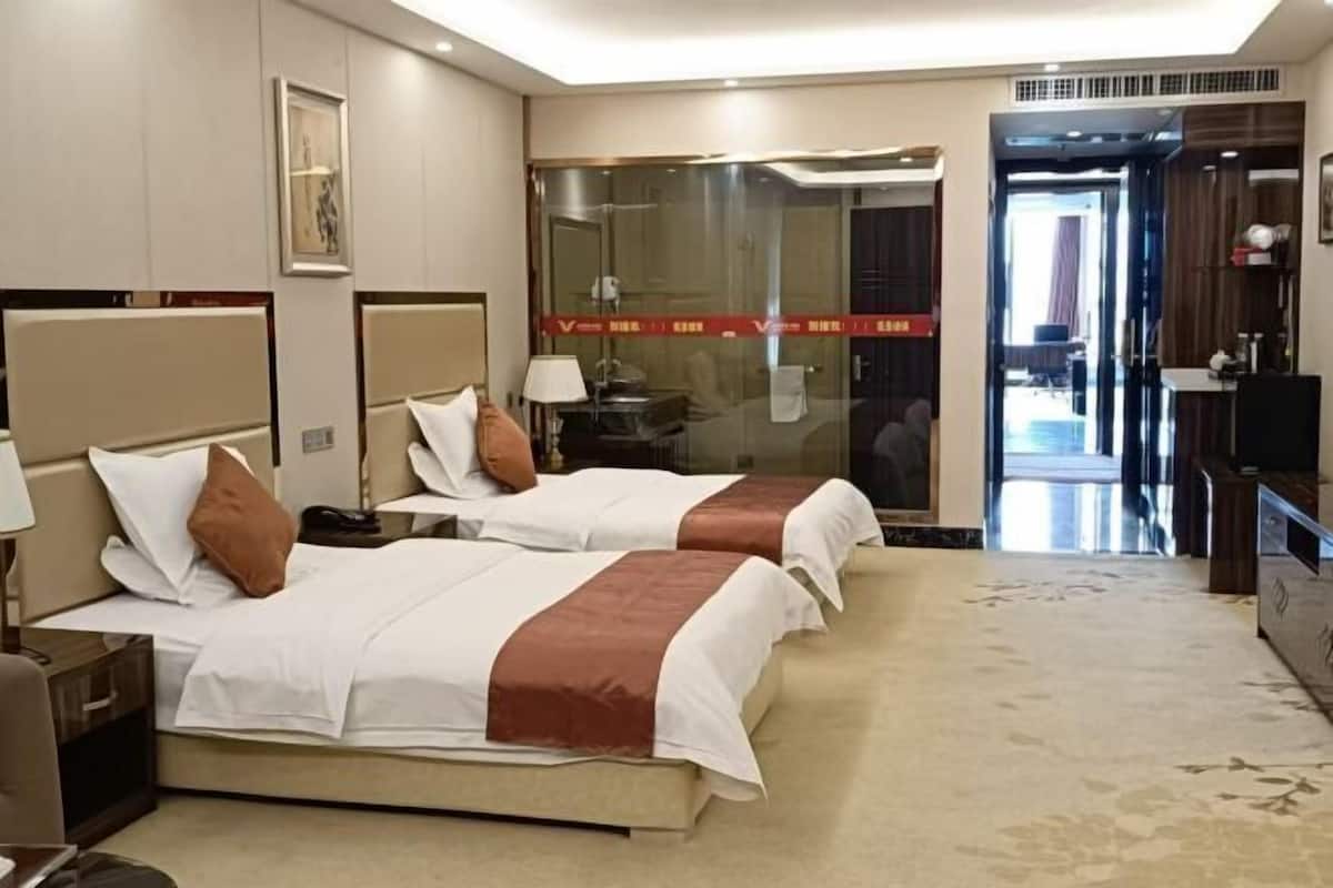 A hotel room with two beds, a desk, a chair, a TV, and a bathroom visible through a glass door.