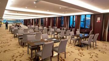 A hotel dining area with multiple tables and chairs, large windows, and a carpeted floor.