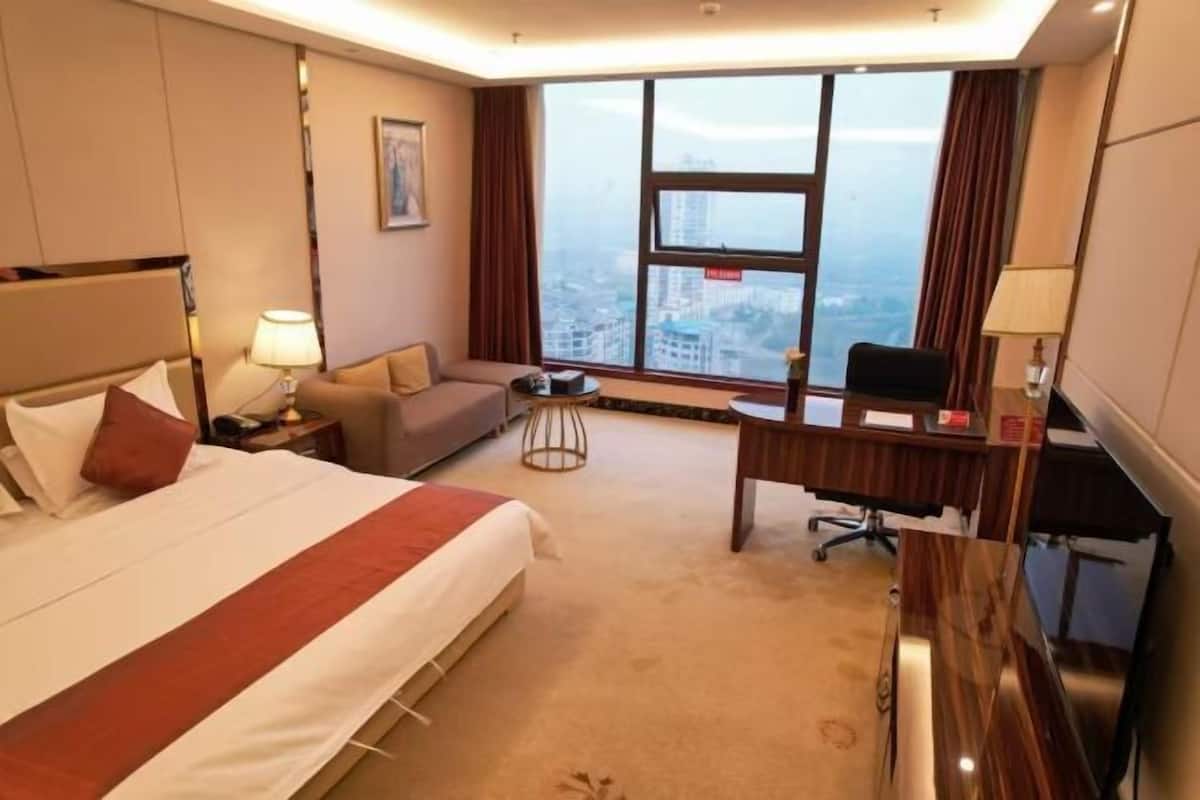 A hotel room with a bed, a desk, a sofa, a window with a city view, and a TV.