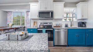 Una cocina moderna con armarios blancos y azul verdoso, una encimera de granito, electrodomésticos de acero inoxidable y una zona de comedor con mesa y sillas.