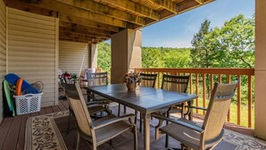 Eine Terrasse im Freien mit Holztisch und -stühlen, einem Korb voller Gegenstände und Blick auf Bäume.