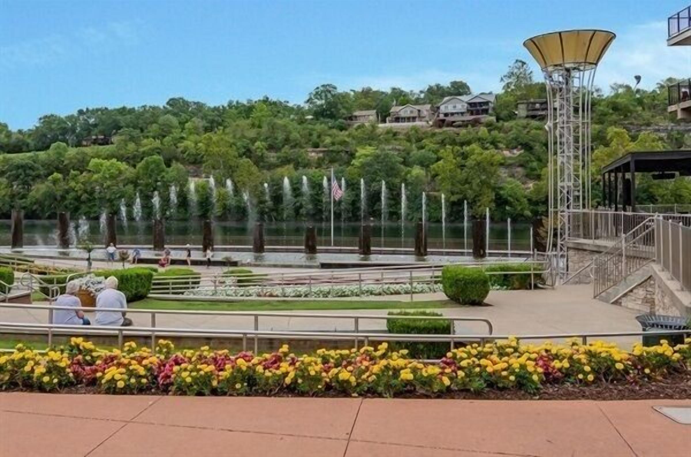 A landscaped area with a fountain, a tall structure with a yellow canopy, and a view of houses on a hill.