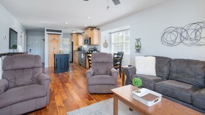 Amplia sala de estar con un sofá gris, dos sillones y una mesa de centro de madera. Se ve un ventilador de techo y una cocina al fondo.