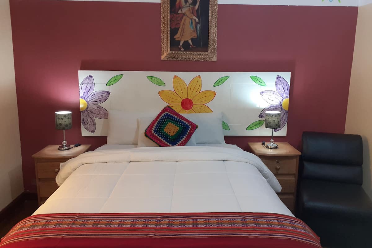 Ein Hotelzimmer mit einem Bett, zwei Nachttischen, einem Sessel sowie einer Wand mit floralen und religiösen Bildern und einem gerahmten Bild über dem Bett.