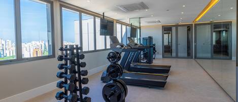 A modern gym with treadmills, dumbbells, and a rowing machine, large windows with a city view, and a glass wall reflecting the interior.