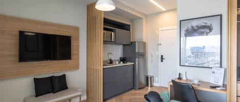 Ein modernes Hotelzimmer mit Küchenzeile, einem Flachbildfernseher an der Wand, einer Bank, einem Sofa und einem Schreibtisch mit Tasse und Speisekarte.