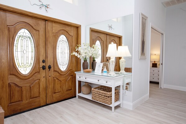 Una habitación con puerta de madera y paneles de vidrio, una mesita consola blanca con un jarrón de flores y un espejo encima.