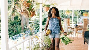 Un espacio al aire libre techado con plantas colgantes, muebles de mimbre y una persona sosteniendo un coco y plátanos.