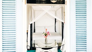 Una habitación con una cama con dosel, una mesa de vidrio con un florero y una silla blanca con cojín azul turquesa.