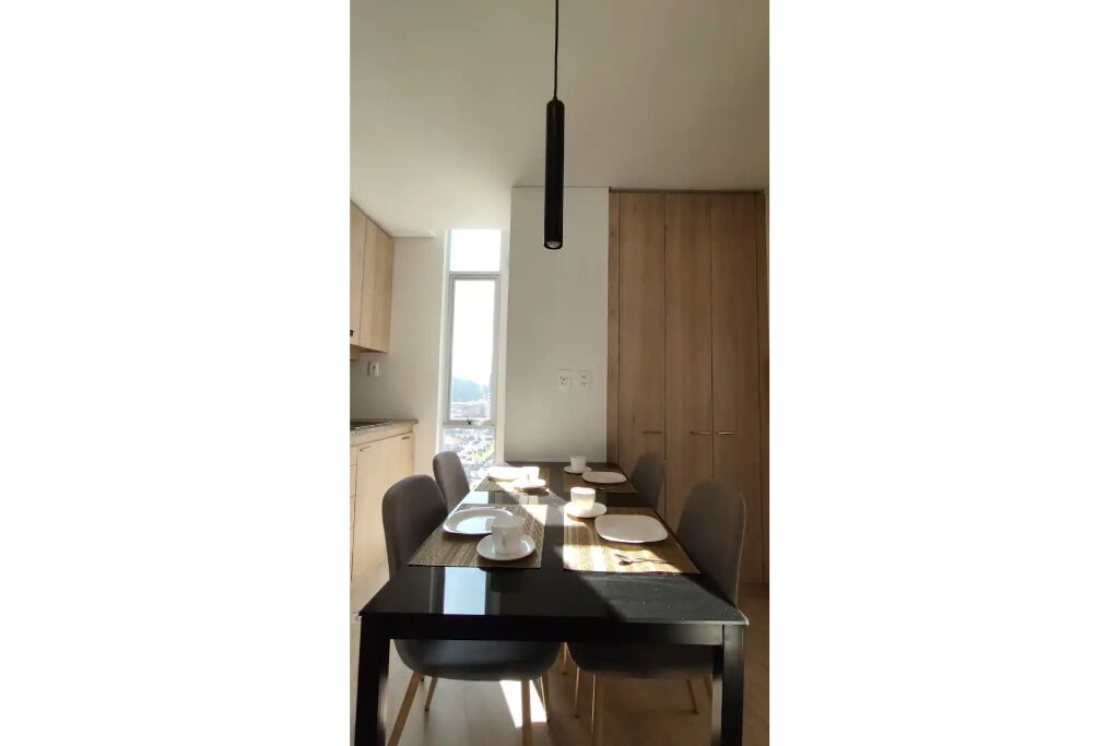 A modern dining area with a black table and chairs, a hanging pendant light, and a view of the city through a large window.