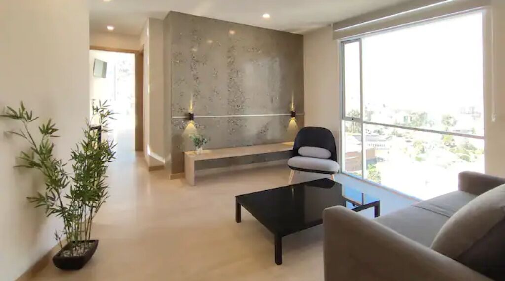 A modern living room with a sofa, a coffee table, and a bench. There is a large window providing a view of the outside.