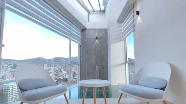 A modern room with two grey chairs and a small table, large windows with blinds, and a city view.