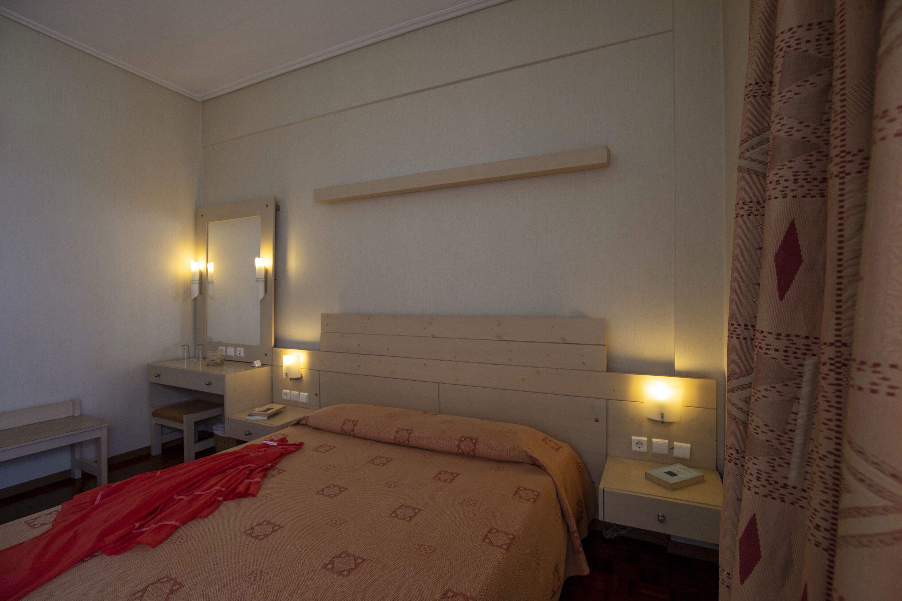 A hotel room with a bed, bedside table, nightstand, mirror, and a wall-mounted shelf.