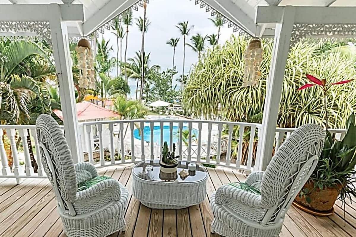 A covered outdoor seating area with wicker chairs and a small table, overlooking a pool and tropical vegetation.