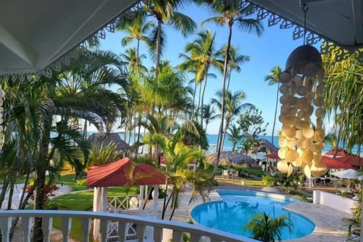 A pool area with a clear view of the ocean, surrounded by palm trees and a thatched-roof structure.
