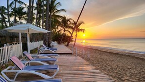 A wooden deck with lounge chairs and a white fence, overlooking a beach at sunset. - Hotel Casa Coson (Las Terrenas)